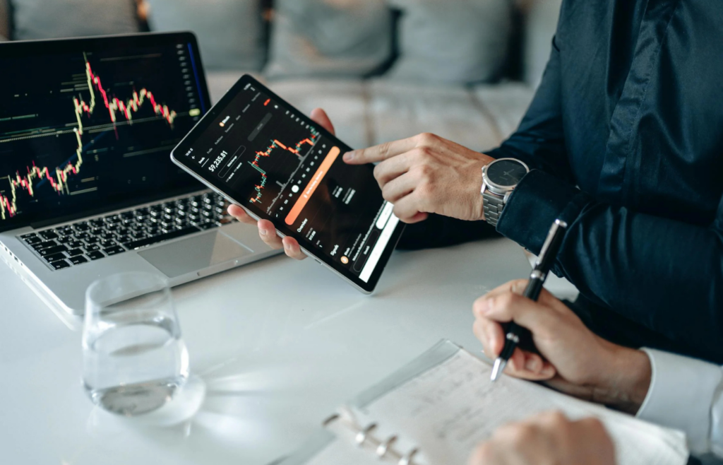 A person looking at a clock next to a laptop with stock market data on the screen