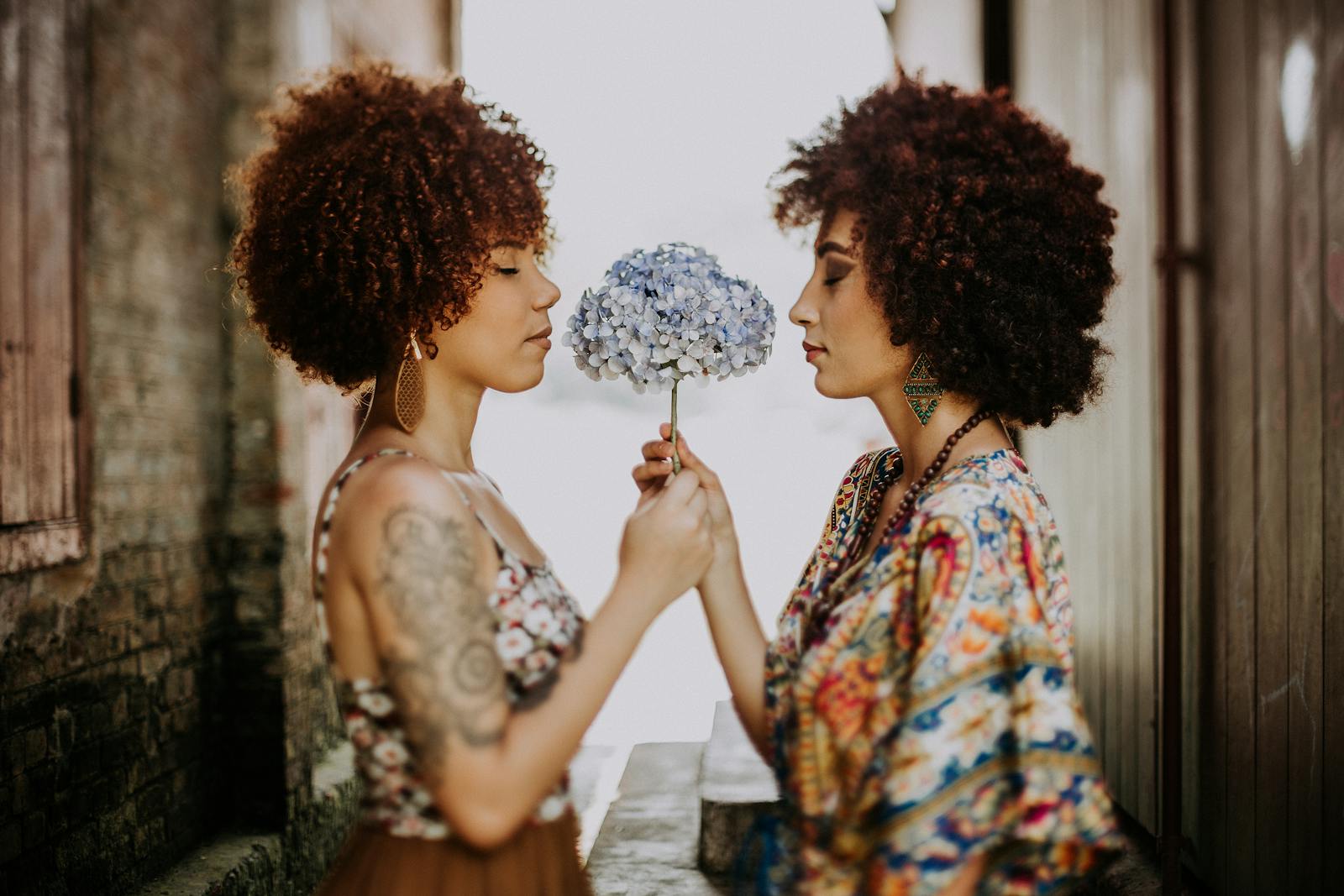 Two fashionable women with curly hair share a moment, holding hydrangea flowers.