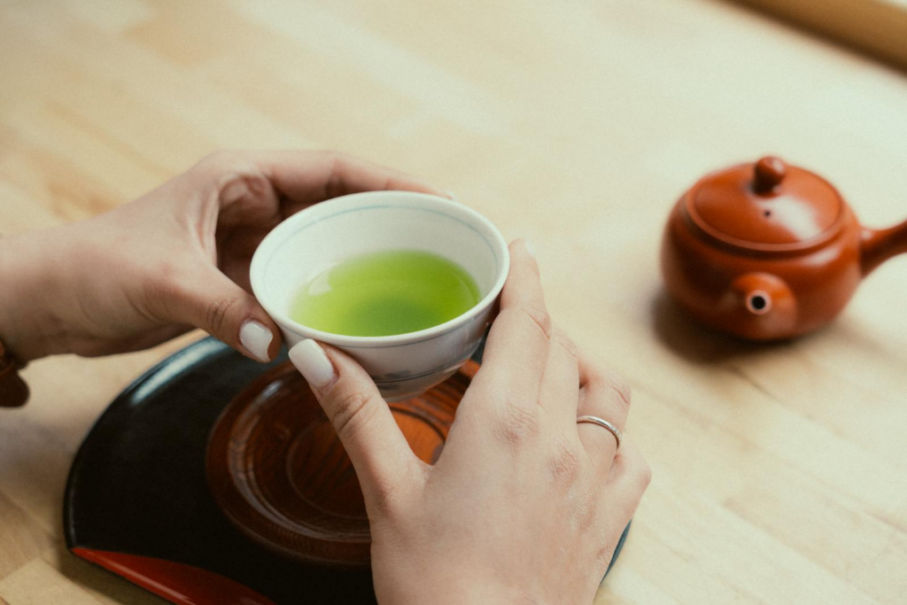 Hands holding a cup of green tea next to a traditional Japanese teapot.