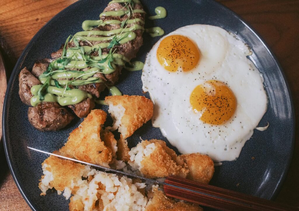 Delicious breakfast plate featuring steak with sauce, fried eggs, and crispy potato cakes.