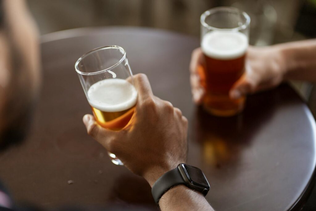 Two individuals enjoying alcoholic beverages in a casual setting, focus on hands and beer glasses.