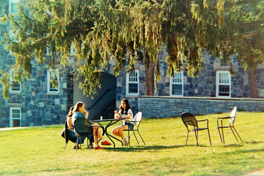 Two women sit at a table outside a stone building.