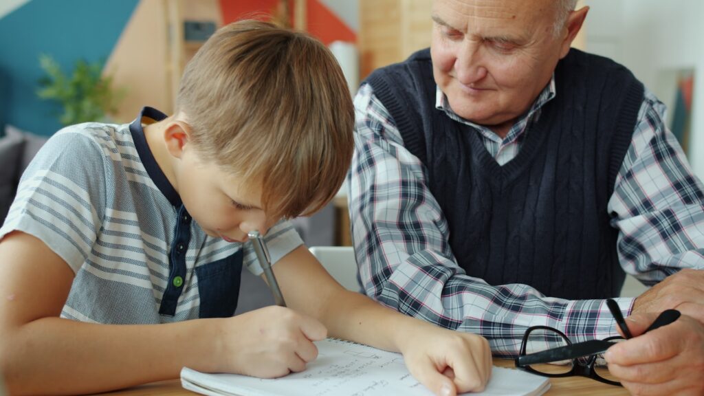 Grandfather helps grandson with homework at table.