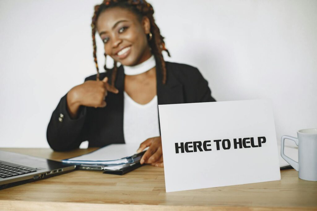 Smiling businesswoman ready to assist, sitting at desk with helpful sign.