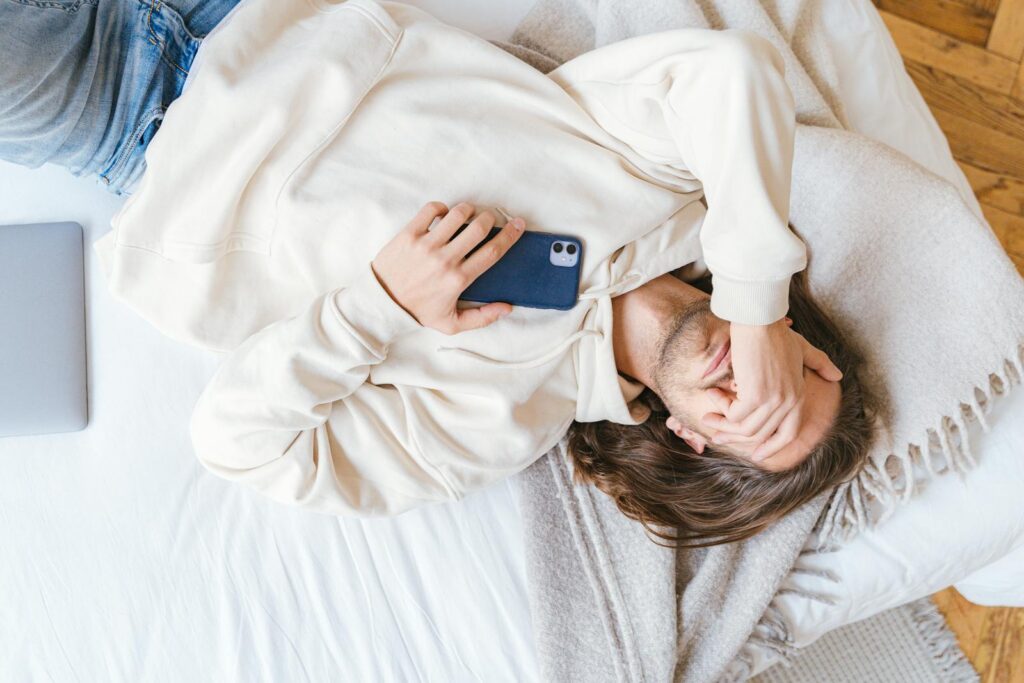 A man in a beige sweater holding a smartphone while lying on a bed indoors.