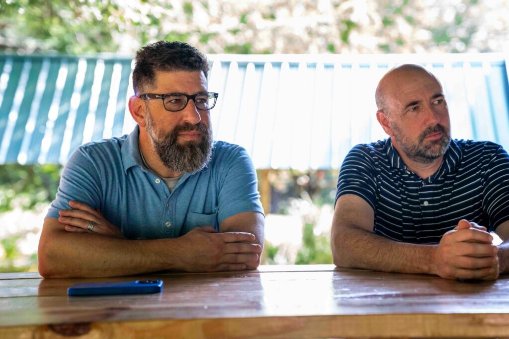 Two men sitting at a wooden table outdoors.