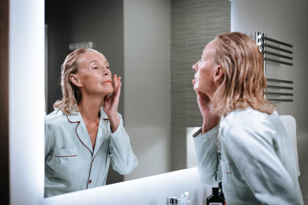Elderly woman admiring her skincare results in a well-lit bathroom mirror.