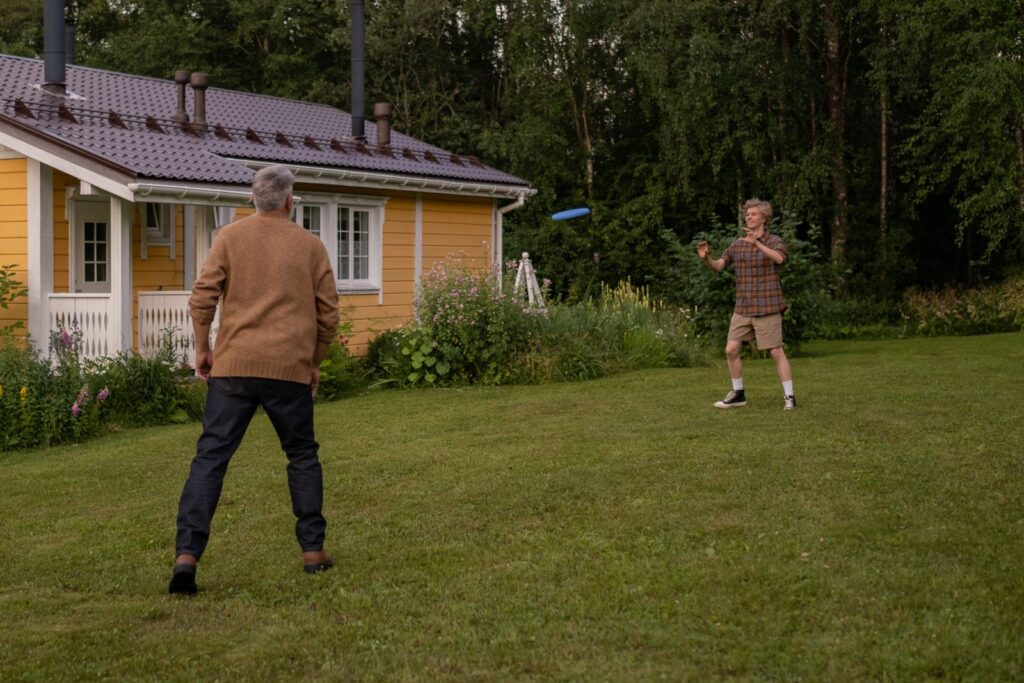 An elderly man and teenager enjoy a frisbee game near a countryside home.