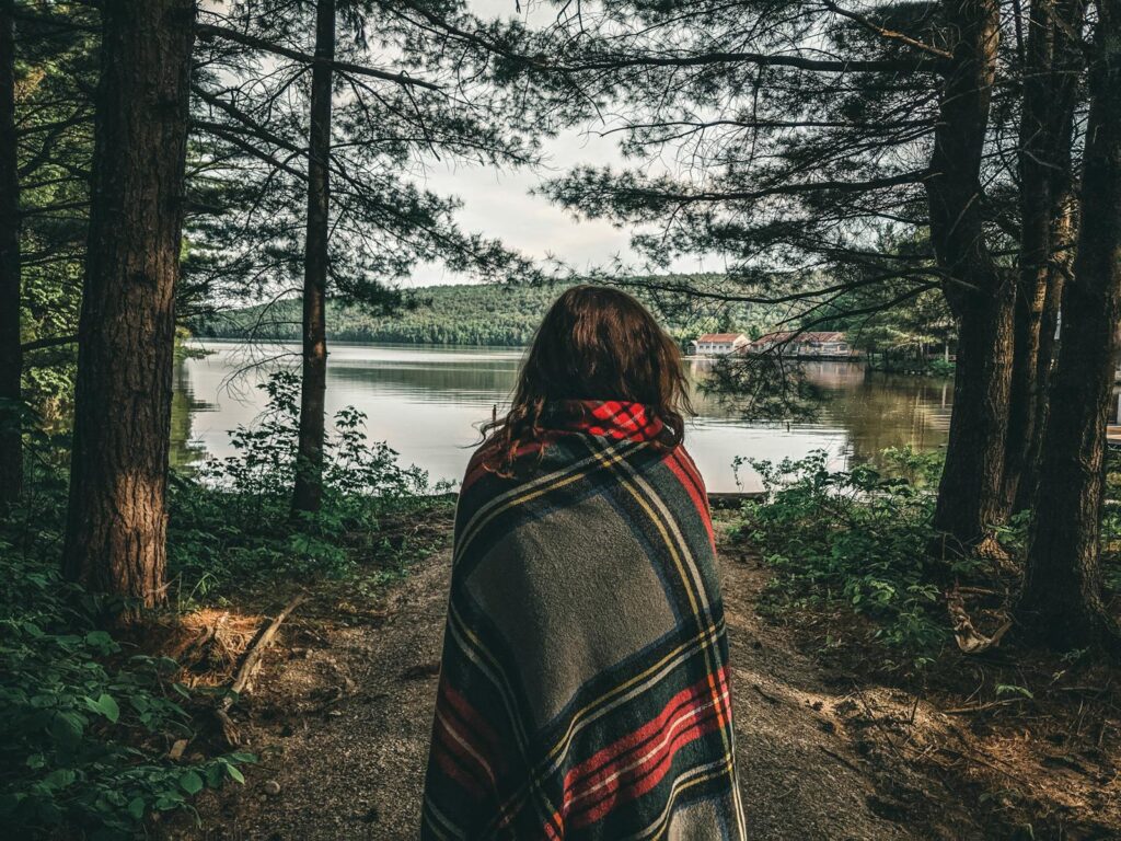 A woman wrapped in a blanket stands in a tranquil Canadian forest by a lake.