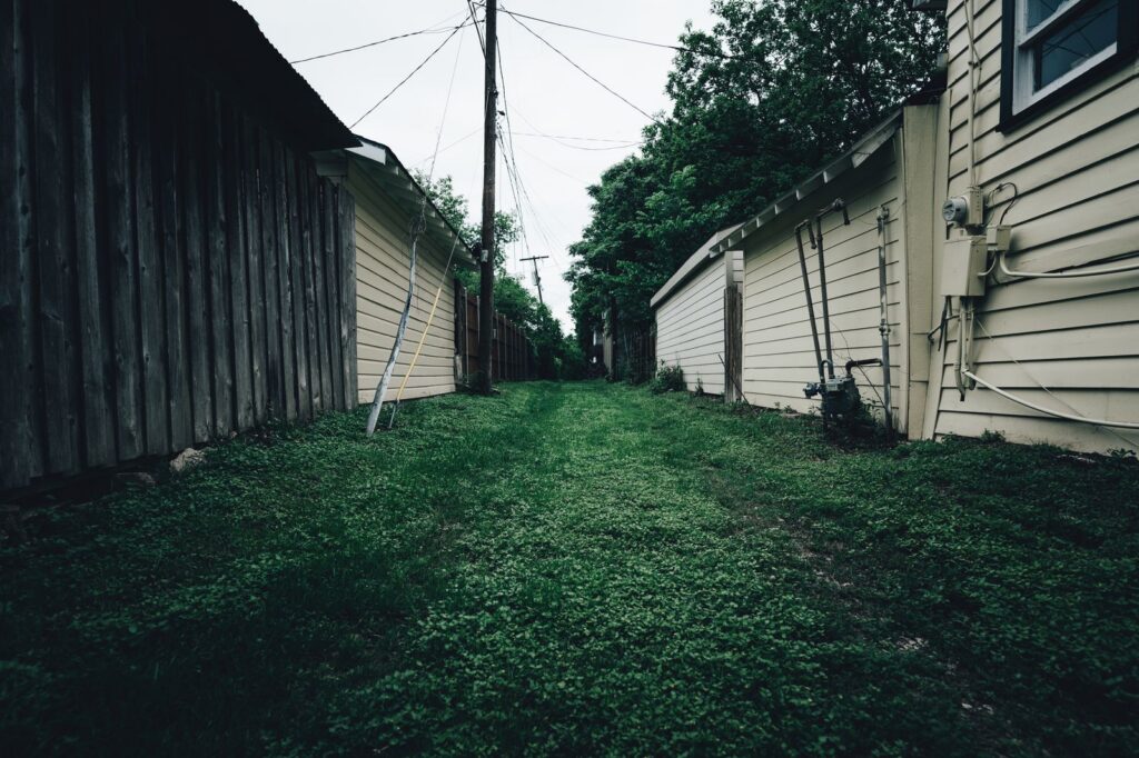 Rustic alleyway with grass pathway between wooden and metal fences in a residential area.
