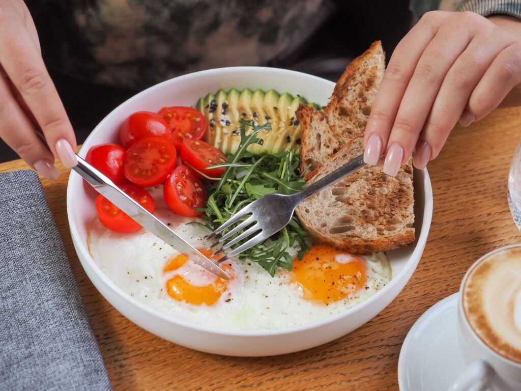 Close-up of a breakfast bowl with fried eggs, avocado, tomatoes, and bread, served with cappuccino.