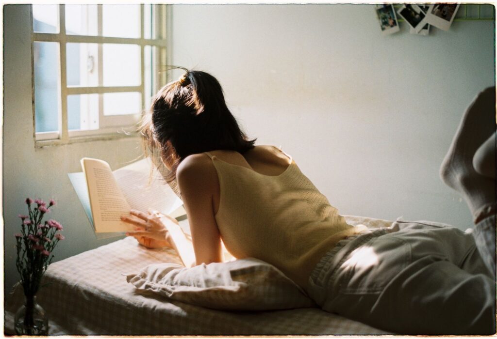 A woman lies on her stomach reading a book by the window, enjoying a peaceful afternoon.