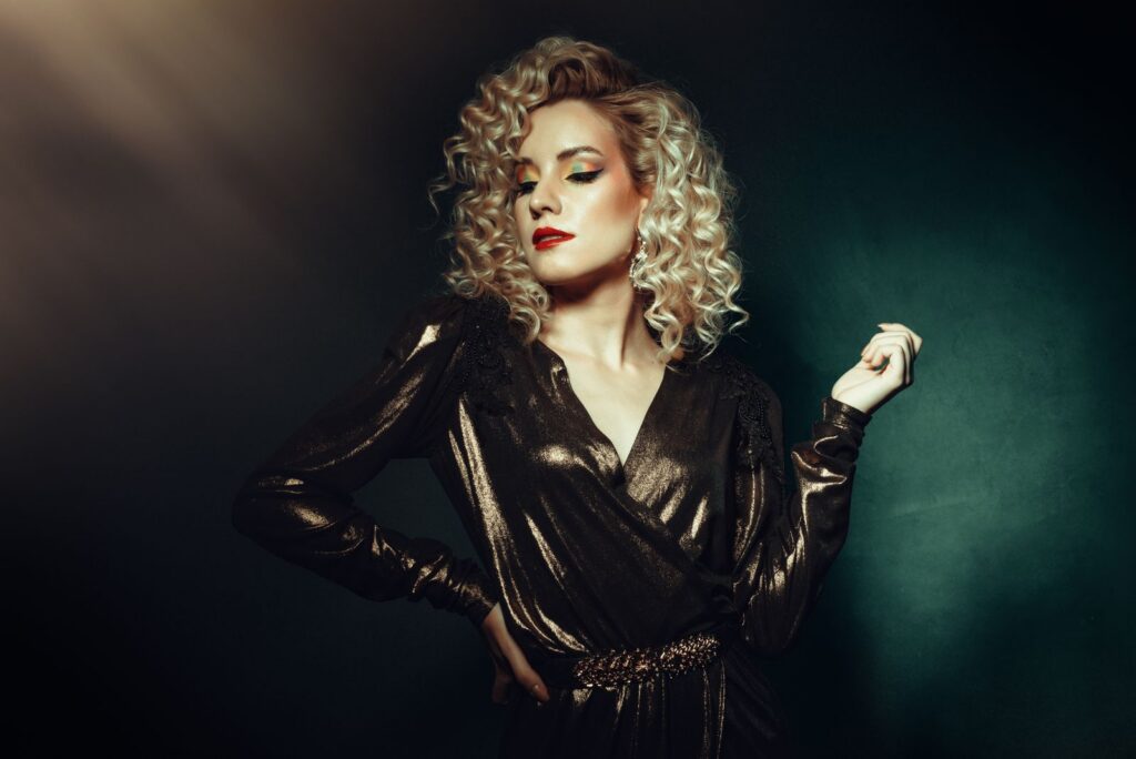 Glamorous blonde woman posing confidently in elegant evening wear with dramatic lighting.