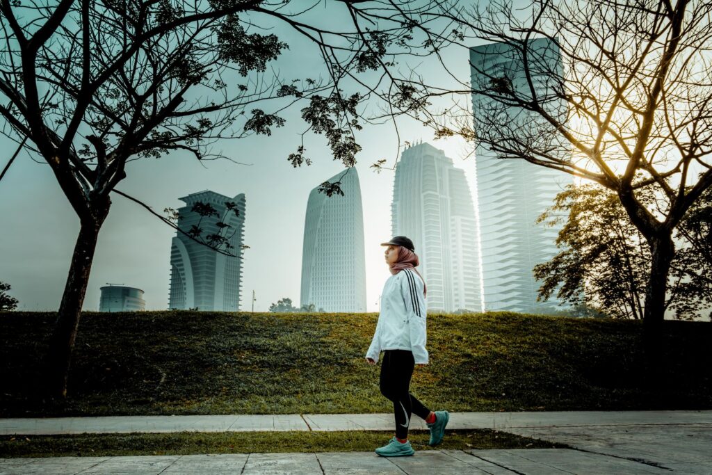 A woman walks in a Putrajaya park, framed by trees with urban high-rises in the background.