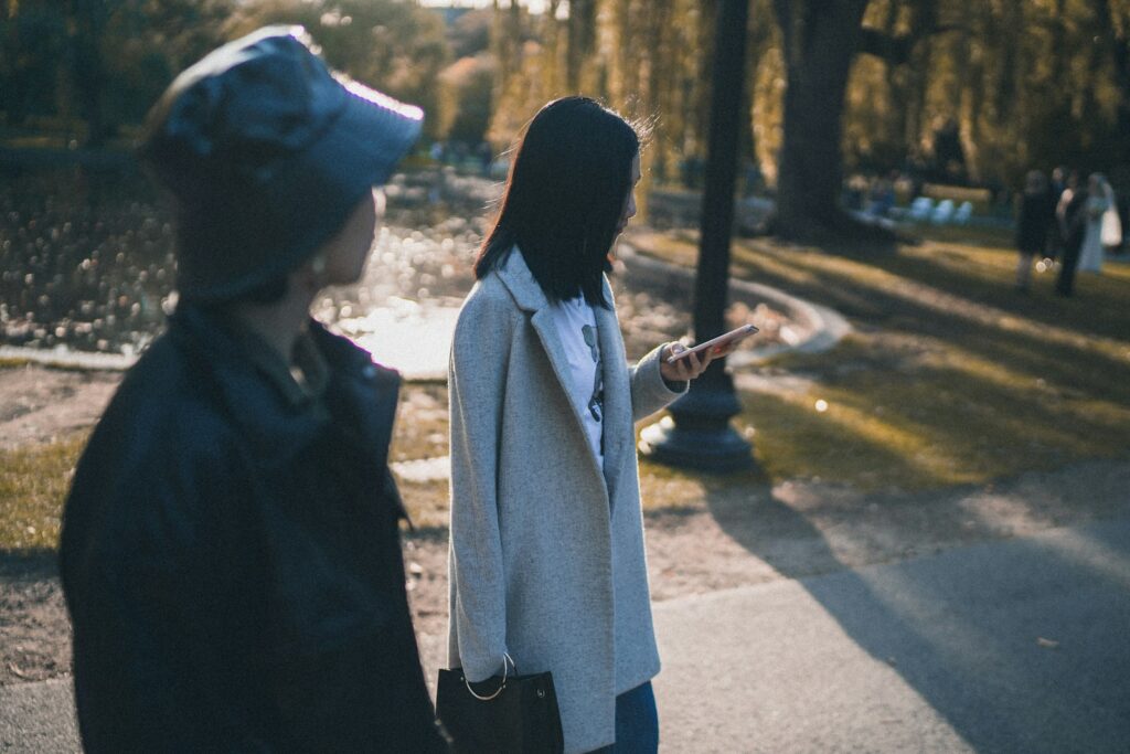woman in white coat holding smartphone standing on sidewalk during daytime