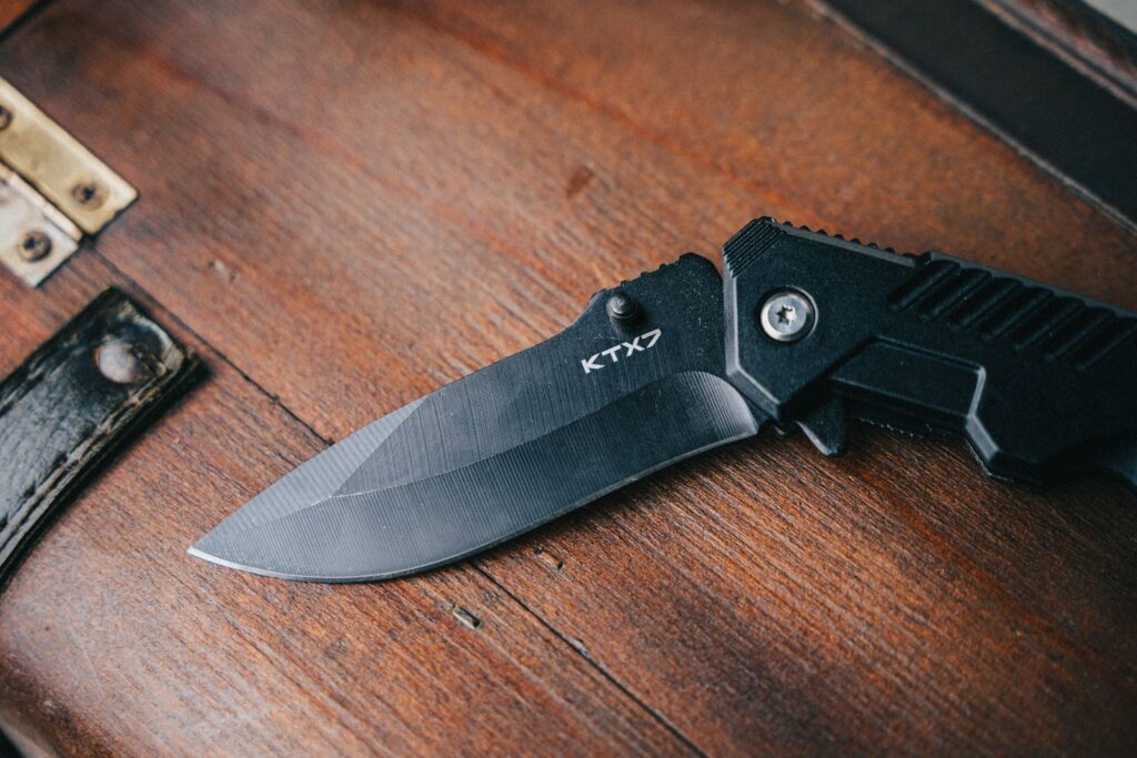 Close-up of a black folding knife on a textured wooden surface showing its sharp blade.