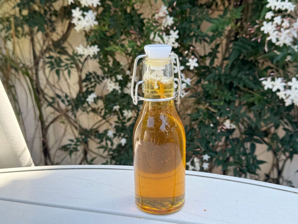 A glass bottle of olive oil placed on a table outdoors with white flowers in the background.