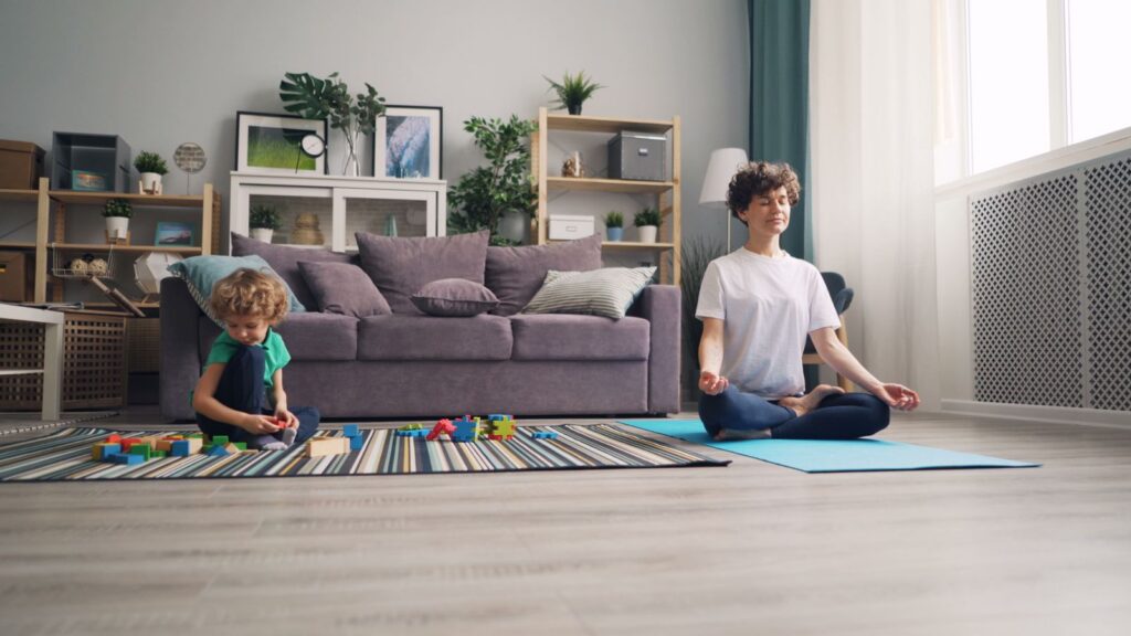 A mother meditates on a yoga mat while her son plays with blocks in a cozy living room setting.