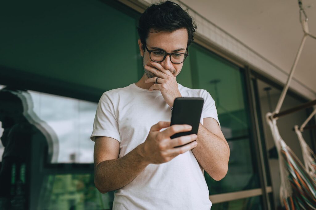 A young man in glasses standing on a balcony, using his smartphone with a thoughtful expression.