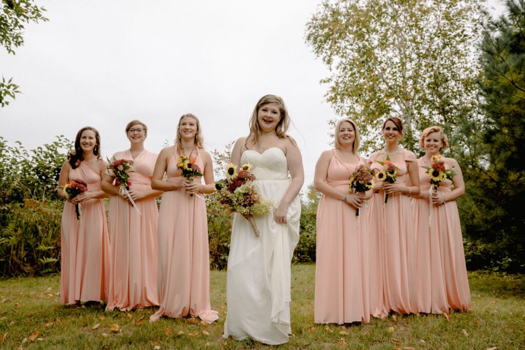A joyful bride with her bridesmaids in a lush outdoor setting, dressed in elegant light pink dresses.