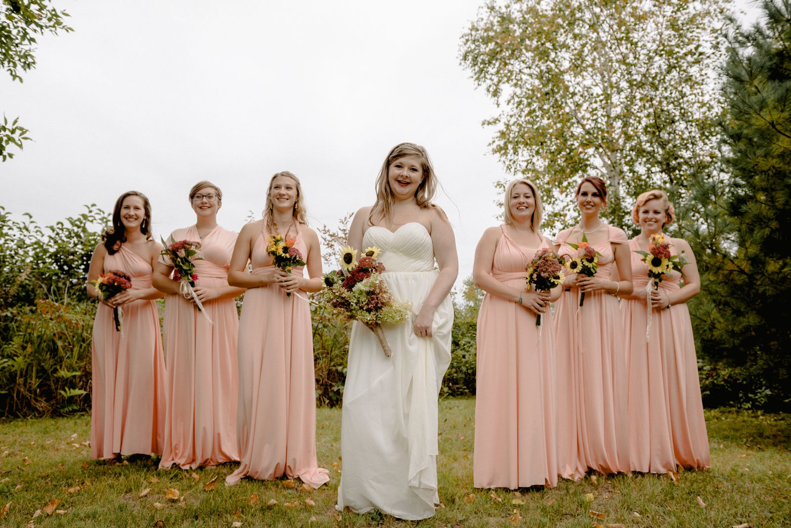 A joyful bride with her bridesmaids in a lush outdoor setting, dressed in elegant light pink dresses.