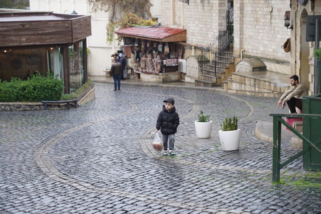 A boy standing on a cobblestone street near an outdoor market, holding a plastic bag in a quaint setting.