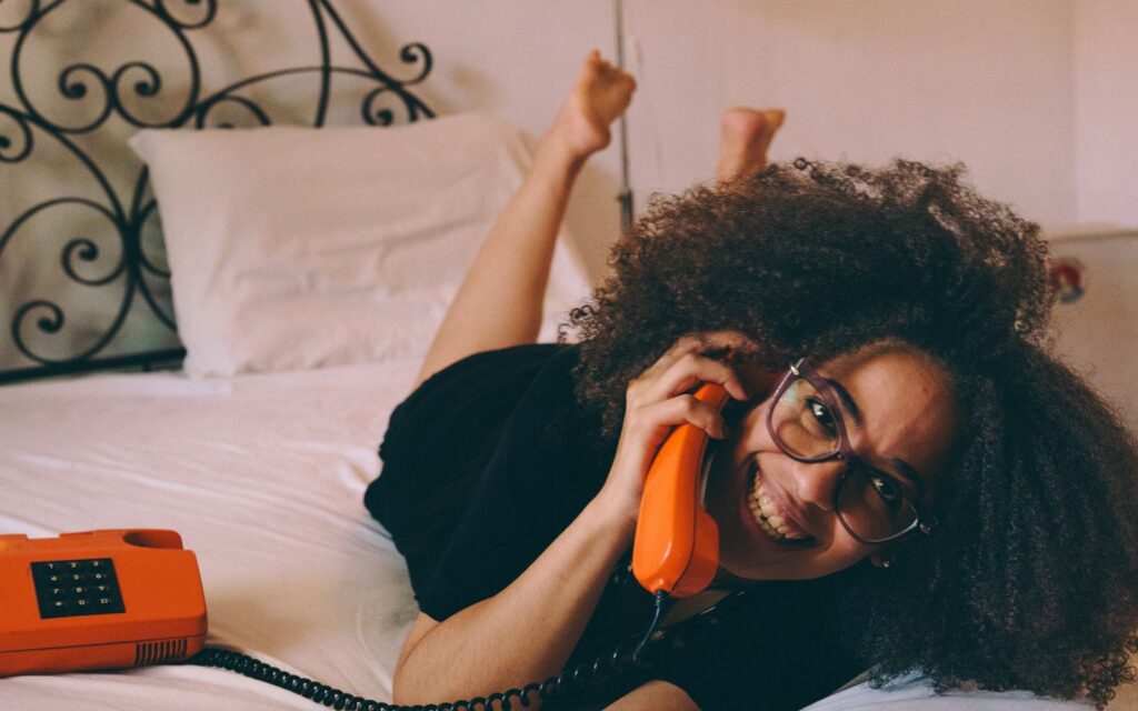 Smiling girl with curly hair chatting on a vintage orange phone while lying on a white bed.