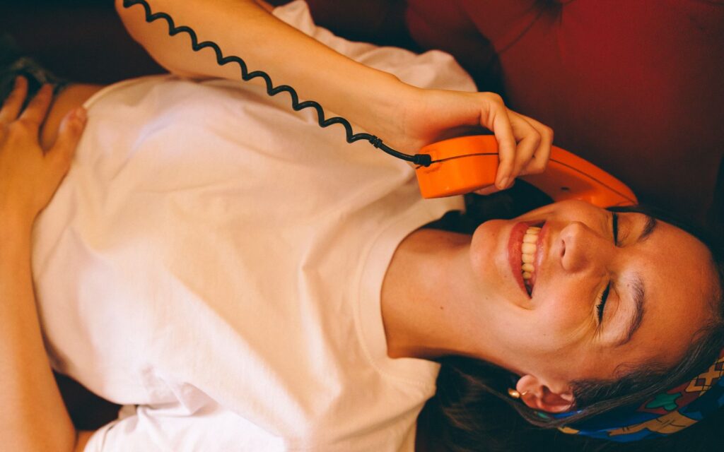 A joyful woman wearing a white t-shirt smiles while chatting on a vibrant orange retro phone indoors.