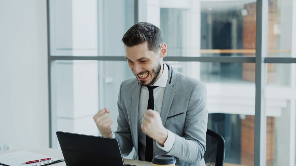 Man in grey suit celebrating success at his desk with a laptop.