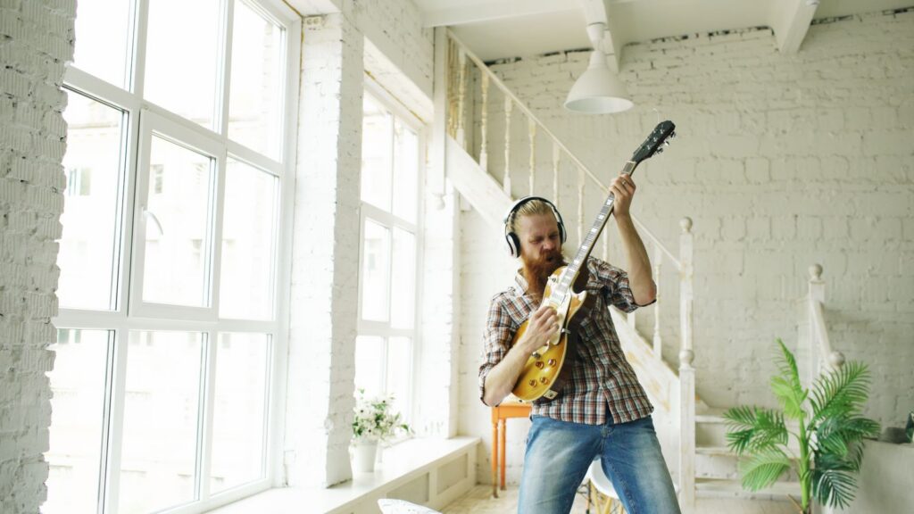 Bearded man wearing headphones passionately plays electric guitar in a bright, modern room.