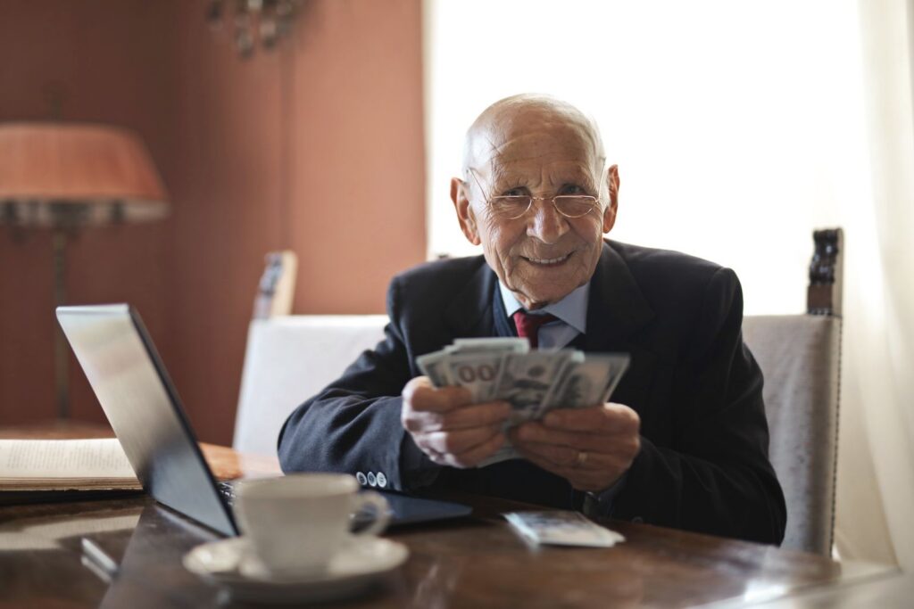 Positive senior businessman in formal suit and eyeglasses holding money in hands while sitting at table near laptop and looking at camera