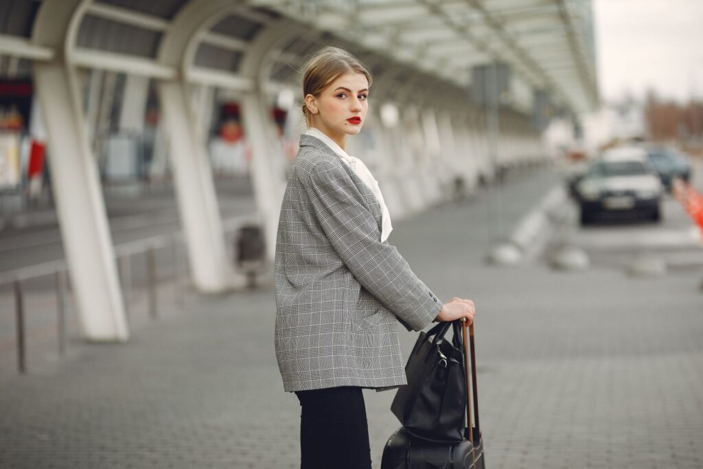 Serious female employee in elegant jacket looking at camera while standing with luggage in airport terminal on street
