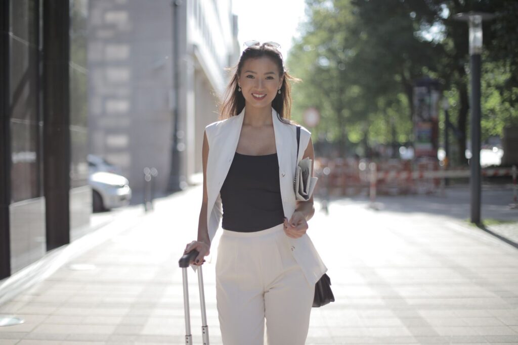 Confident woman in urban setting, carrying luggage and a newspaper, smiling on a sunny day.