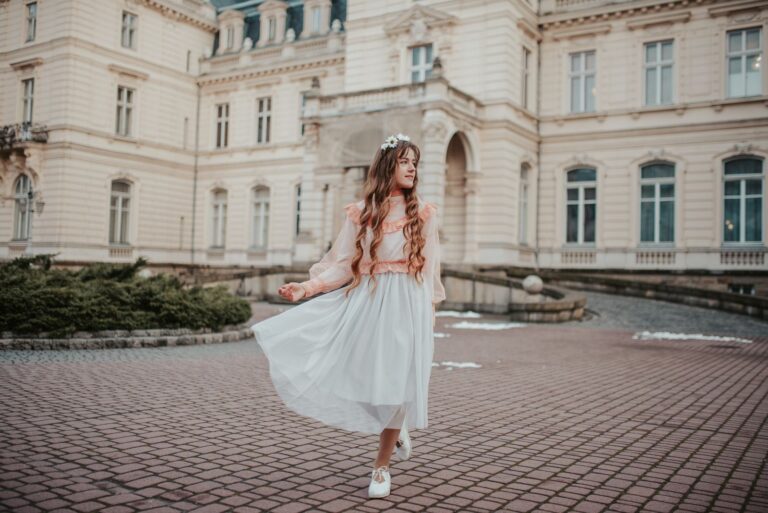 A young woman in a flowing white dress stands gracefully in front of a historic building, exuding elegance and style.