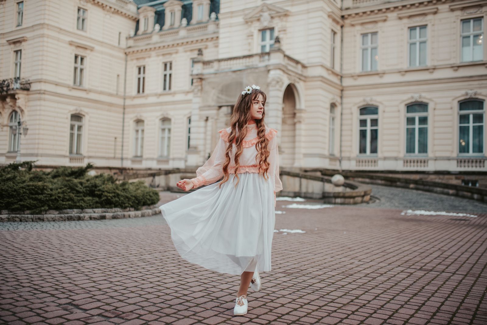 A young woman in a flowing white dress stands gracefully in front of a historic building, exuding elegance and style.