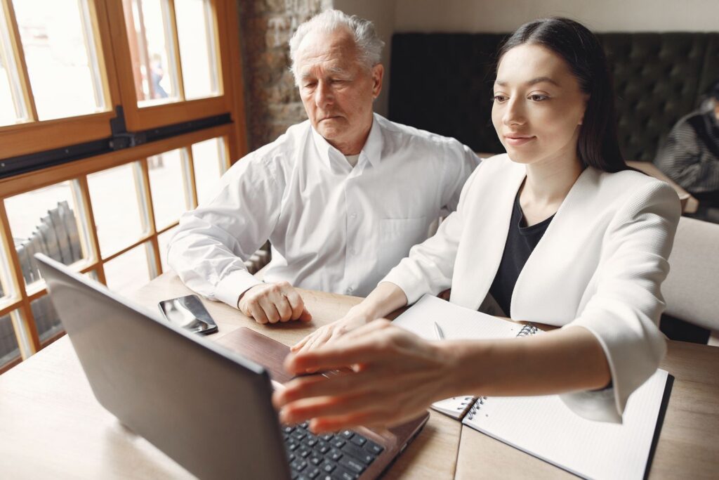Modern adult woman with senior male executive browsing laptop and checking notes while working remotely in creative workspace near window and looking at screen