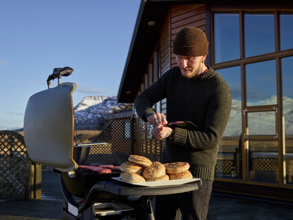 Concentrated young male in warm outfit preparing delicious grilled meat while standing against cozy house in winter day