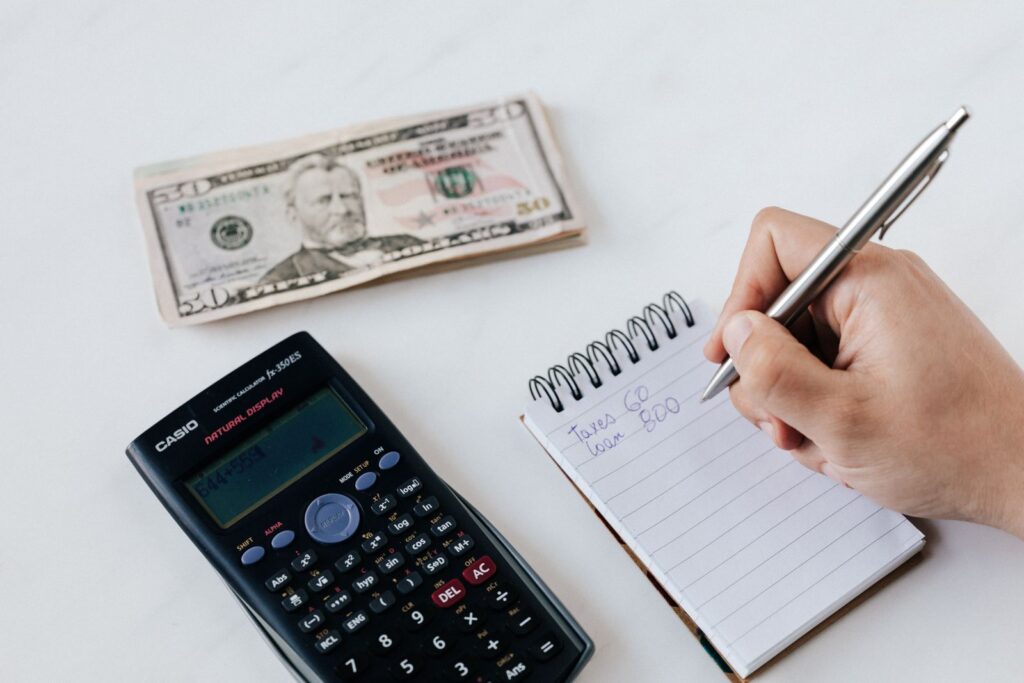 Top view of crop faceless accountant using pen and notepad while counting expenses on table near pile of paper money and calculator