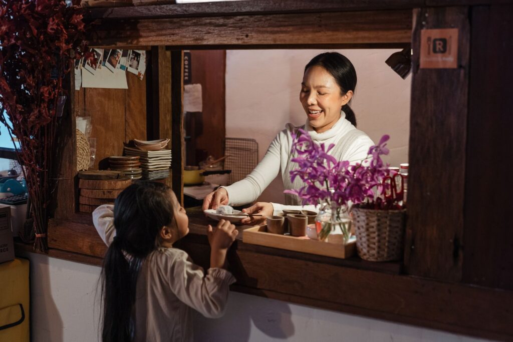 Smiling Asian female cafe owner in apron standing behind wooden counter in kitchen while giving tasty pastry to little girl customer