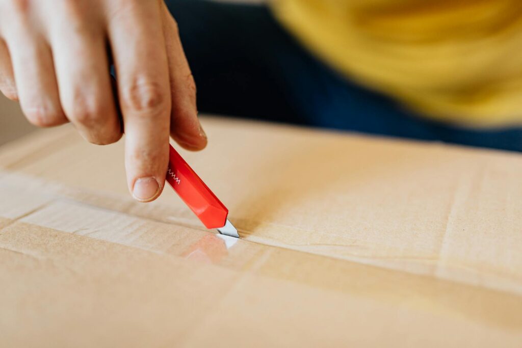 Crop anonymous male unpacking belongings from cardboard box by using sharp utility knife after getting delivery during relocation