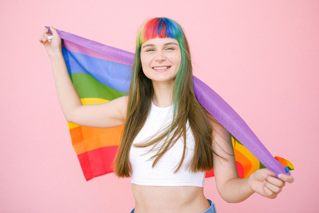 Smiling young woman with rainbow hair holding a pride flag against a pink backdrop, celebrating LGBTQ pride.