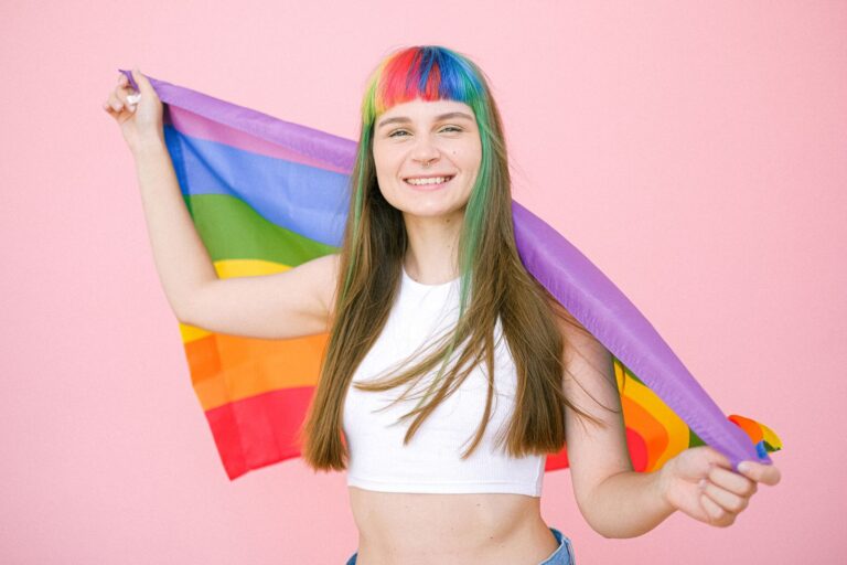 Smiling young woman with rainbow hair holding a pride flag against a pink backdrop, celebrating LGBTQ pride.