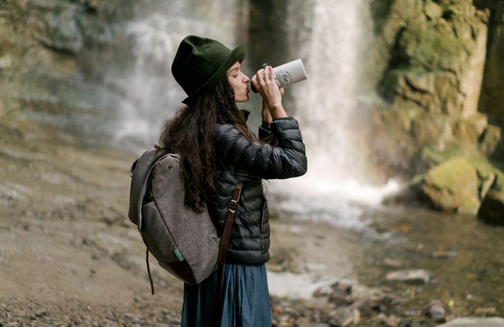A woman drinks from a tumbler while hiking near a waterfall, emphasizing hydration and outdoor adventure.