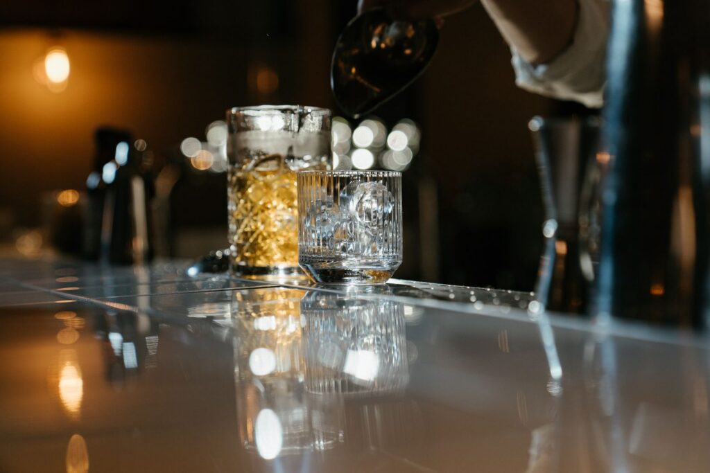Bartender pouring a drink into a glass with ice cubes at a dimly lit bar.