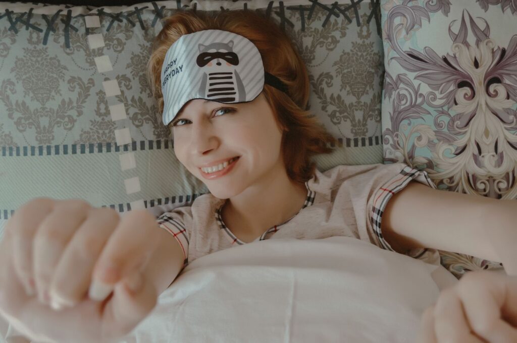 Top view of smiling female reaching out hands to camera while lying on bed under blanket and looking at camera