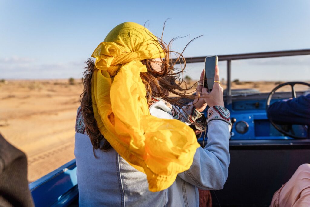 A woman with a yellow scarf captures a desert scene on a safari adventure. Vibrant and adventurous.