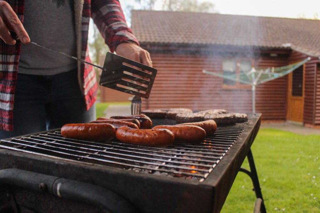 Grilling sausages and patties in a backyard with a rustic wooden house in the background.