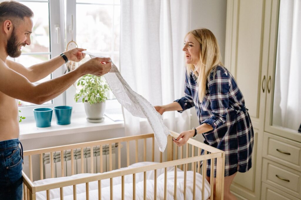 A smiling couple sets up a crib in a sun-filled room, preparing for a new baby's arrival.