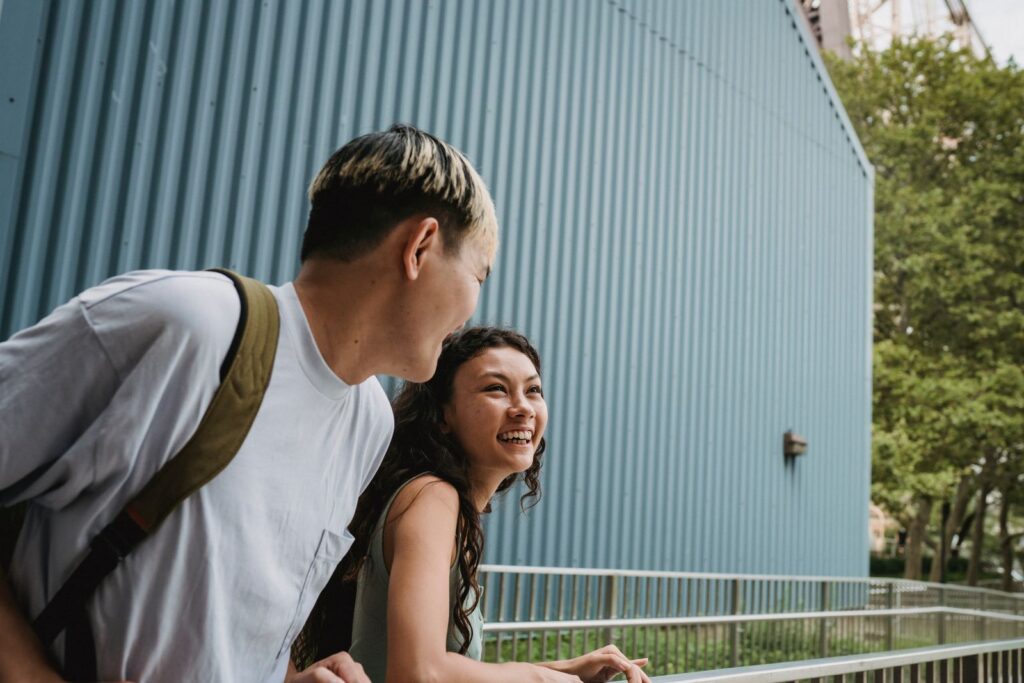 Cheerful ethnic woman speaking with unrecognizable Asian boyfriend while spending time together near fence in town