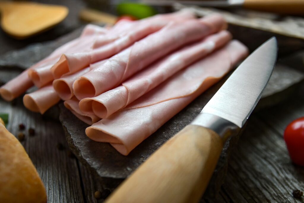 Close-up image of freshly sliced ham with a wooden-handled knife on a slate and wooden table, ready to serve.